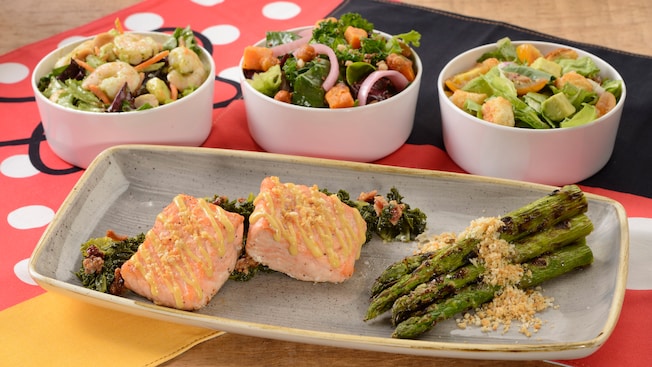 El plato principal de salmón asado con un acompañante de espárragos y diversas ensaladas, incluyendo la ensalada de camarones pequeños, la de temporada y la César