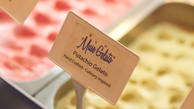 Un contenedor de metal lleno de helado en una caja conservadora y un cartel que dice Pistachio Gelato, echo a mano con motivos culinarios
