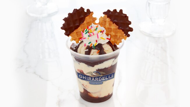 A plastic Ghirardelli cup holds ice cream, hot fudge, whipped cream, sprinkles and 2 chocolate dipped waffle cookies