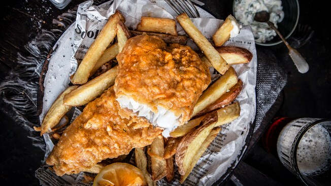 A plate of fish and chips rests on a table alongside utensils, a beverage, a salt shaker and a small bowl of tartar sauce