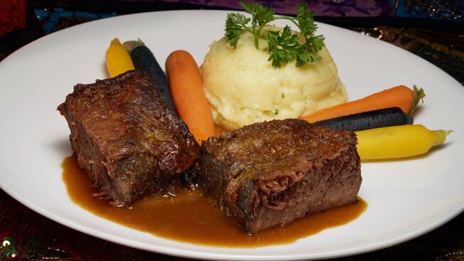 2 braised beef short ribs on a plate with baby carrots and mashed potatoes garnished with cilantro