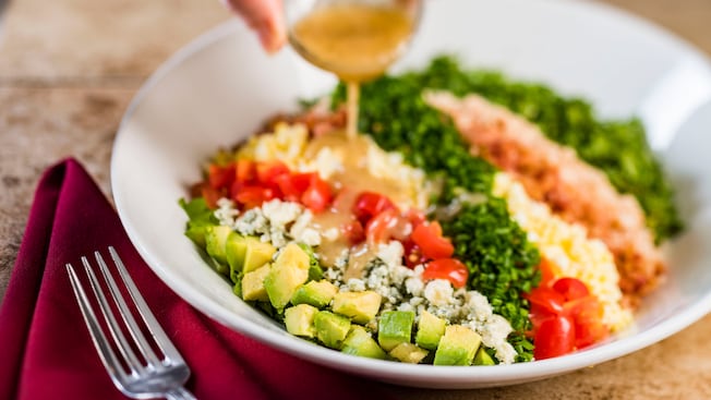 A cobb salad with avocado, bacon, lettuce, tomatoes, and more with a dressing being poured on from a hand above the plate