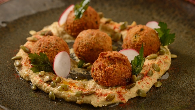 A dish of falafel on top of hummus with sliced radishes and pine nuts