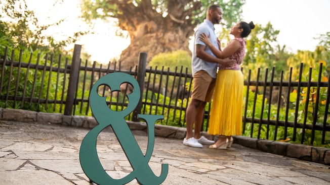 A small ampersand prop with a Mickey Mouse icon inside the top loop rests on the ground. A couple embrace and look into each other’s eyes. The Tree of Life rises behind them