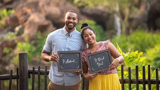 A woman leans into the shoulder of a man. He has a sign that reads I Asked, and she has a sign that reads I Said Yes!