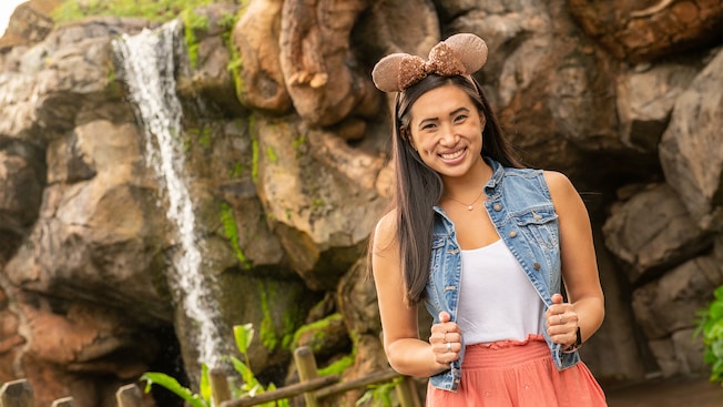 A woman tugs on her jean vest, wears a Minnie Mouse ear hat and poses in front of a waterfall