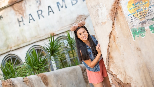 A woman wears a Minnie Mouse ear hat and peaks out from behind a wall, as she stands in front of a weathered building with the word Harambe painted on it