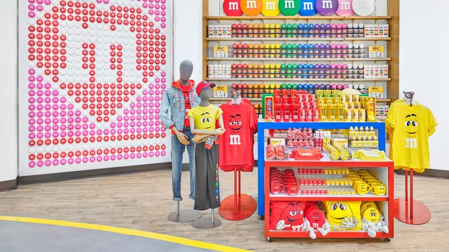 Une grande mosaïque faite de tuiles qui ressemblent à des bonbons M and M, et des présentoirs avec des produits M and M, dont des chandails, des vestes, des tasses, des tasses de voyage, des coussins et des emballages de bonbons
