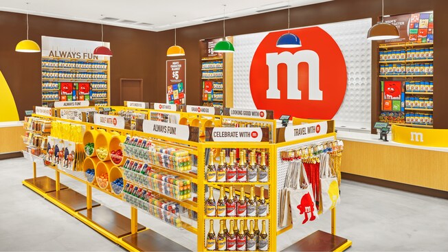 Une caisse et une file bordée de présentoirs avec des produits M and M, dont des sacs fourre-tout, des balles de golf et des bouteilles de champagne remplies de bonbons. Différentes enseignes indiquant Work from Home with M, Character Tote Eco Friendly, Gold with M, Always Fun, Celebrate with M, Travel with M, Looking Good with M, Grab M and Go, Must Haves, Gifts Under $10, et M and Ms Character Blanket $5.