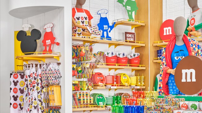 Des présentoirs avec des produits M and M pour la cuisine, dont des bols à mélanger, des tasses à mesurer, des assiettes, des bols, des tasses de voyage, des fouets, des gants de cuisine, des moules à biscuits, des pailles et des tabliers. Une enseigne indiquant « Home and Baking ».