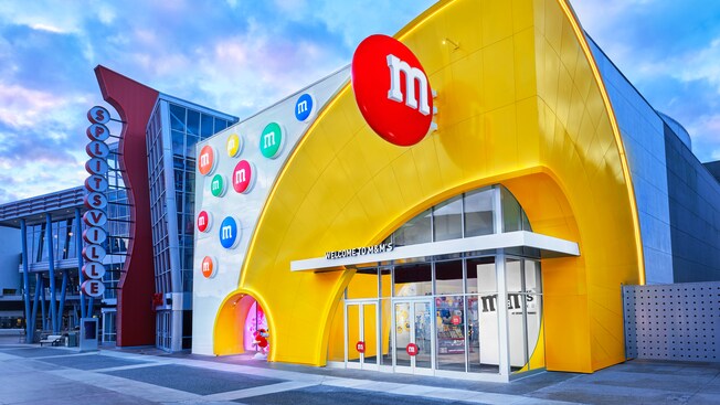 L’extérieur d’un magasin avec une marquise indiquant « Welcome to M and Ms », décoré de bonbons géants M&M, à côté d’un bâtiment avec une enseigne indiquant « Splitsville »