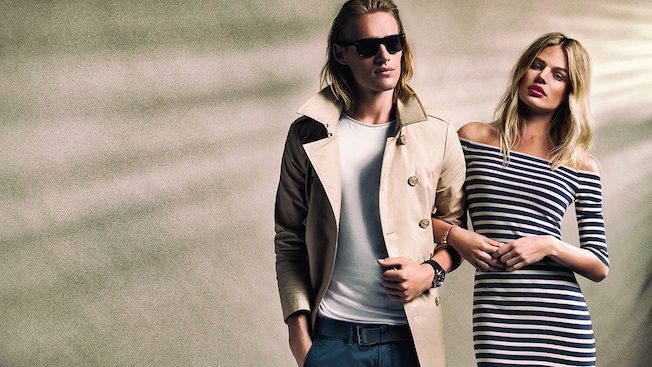A man modeling a trench style jacket and sunglasses alongside a woman modeling an off the shoulder long sleeve dress from Superdry