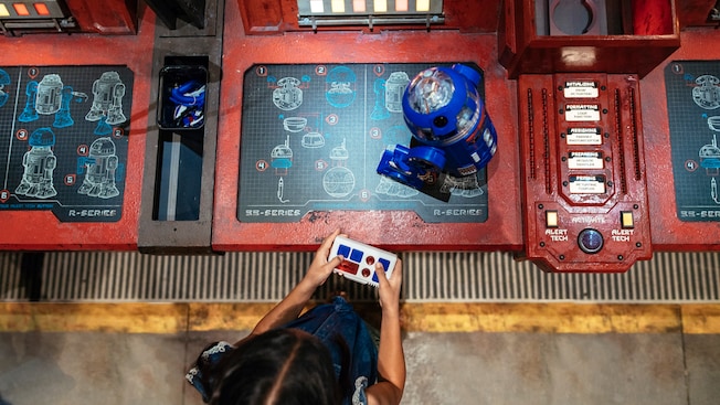 A young girl controls her new R series droid atop a workshop console featuring construction blueprints of droids