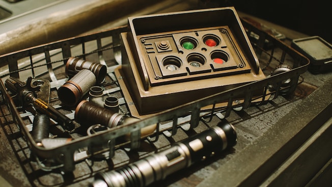 Three images in one include a Savi Gatherer holding a vial containing luminescent kyber crystals, a lightsaber hilt, and a lightsaber hilt besides a carton featuring spare parts