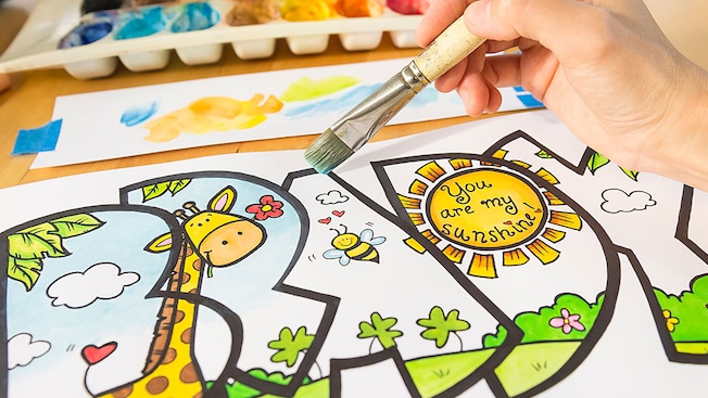 A hand holding a paintbrush, coloring a whimsical meadow scene contained by the outline of the letters of the name Brady