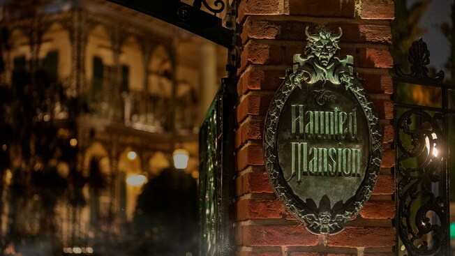 A brick pylon at the entry gate features a sign that reads Haunted Mansion, with the Mansion in the background