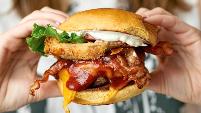 Hands holding a burger with bacon, onion rings and barbecue sauce