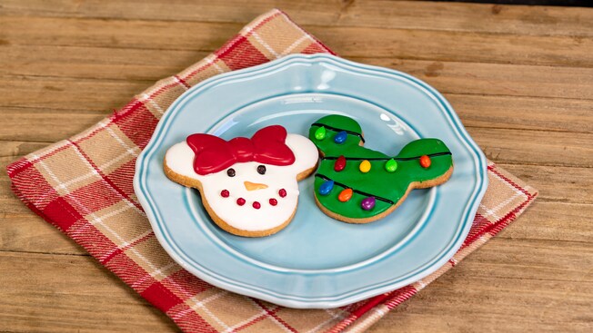 A Mickey ears holiday tree cookie and a Minnie bow snowman face holiday cookie sit on a plate atop a plaid napkin