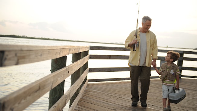 On a pier, a grandfather with a fishing pole holds the hand of his grandson, who holds a tackle box