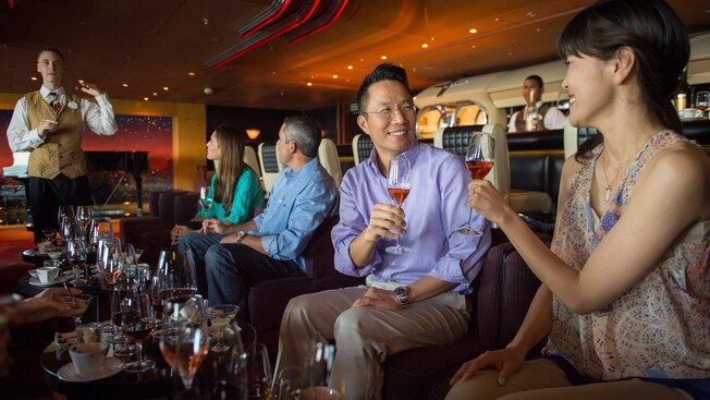 A group of people sit in front of a table full of drinks and glasses as a man speaks to them while holding a drink