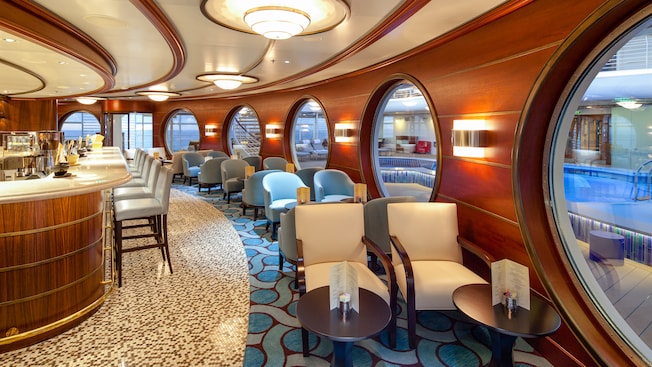 A lounge has high stools at a casual bar and rows of chairs with cocktail tables next to porthole windows A lounge has high stools at a casual bar and rows of chairs with cocktail tables next to porthole windows