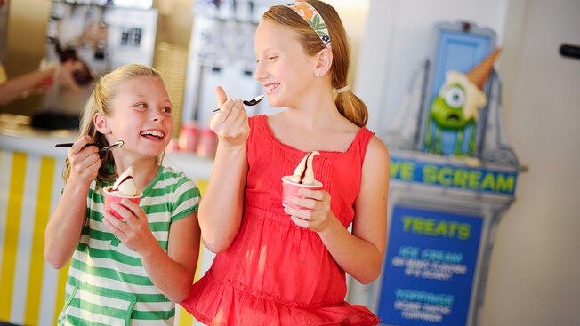Two young girls smile at each other while eating ice cream with chocolate sauce in cups