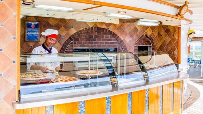 A chef stands behind a display of pizza pies and puts a slice on a plate, at Pinocchios Pizzeria