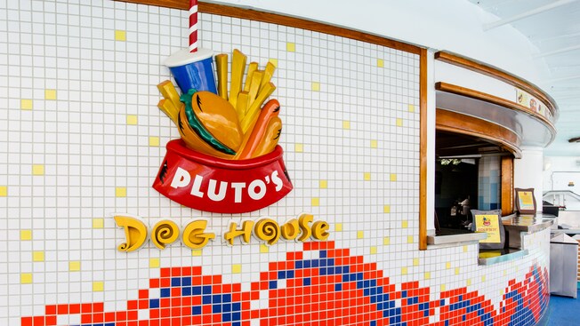 A 3 D sign affixed to mosaic tiles depicts a dog bowl, hamburger and hot dog and says, Plutos Dog House