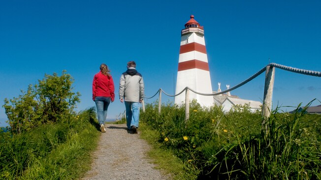 A couple walks along a grassy path toward a lighthouse