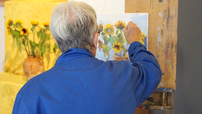A person standing in front of an easel and painting a still life featuring an arrangement of sunflowers