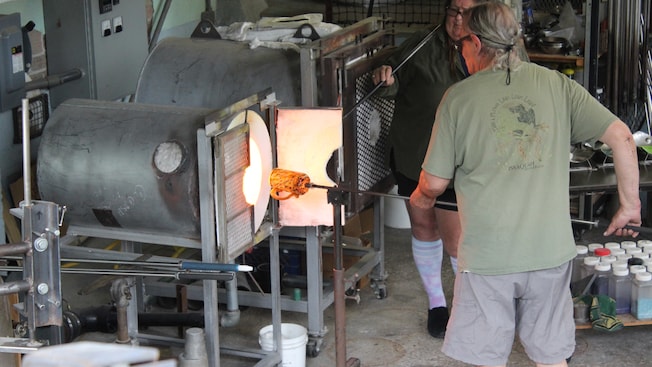 A man fashions a glass mug near a kiln while a woman assists him A man fashions a glass mug near a kiln while a woman assists him