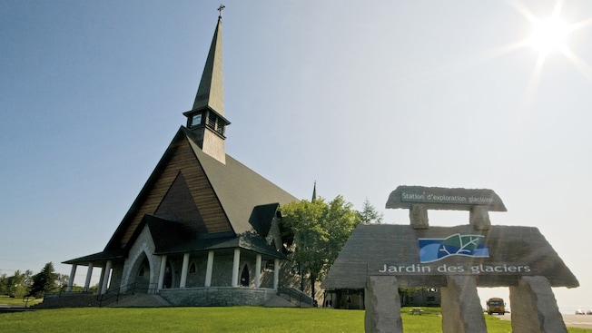 A chapel near trees, a lawn and a sign that reads Jardin Des Glaciers A chapel near trees, a lawn and a sign that reads Jardin Des Glaciers