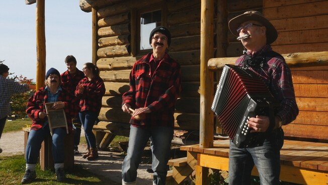 Several musicians play the accordion, harmonica, washboard and other instruments on a log cabin porch