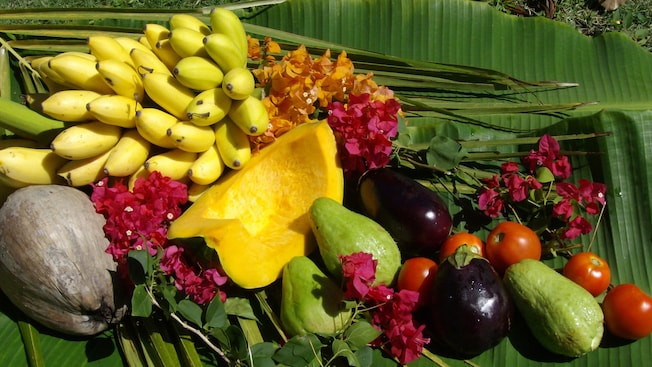 Fresh, whole produce including bananas mangos, tomatoes and a coconut are laid out on a giant palm leaf