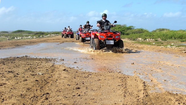 A group of people ride ATVs across a puddle and a dirt road A group of people ride ATVs across a puddle and a dirt road