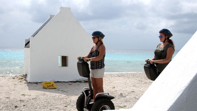 Two women ride Segways on a beach with huts