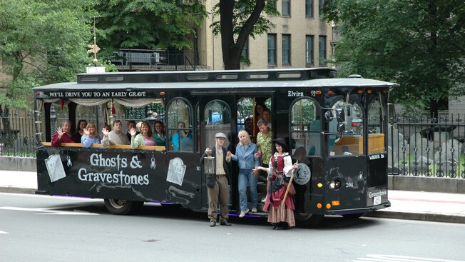 Costumed guides pose in front of a Ghosts & Gravestones themed tour bus as passengers smile and wave