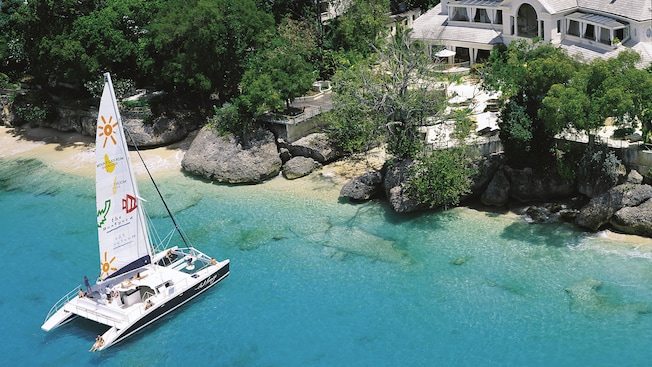 Um catamarã veleiro está muito próximo de uma costa onde um caminho leva a um resort Um catamarã veleiro está muito próximo de uma costa onde um caminho leva a um resort