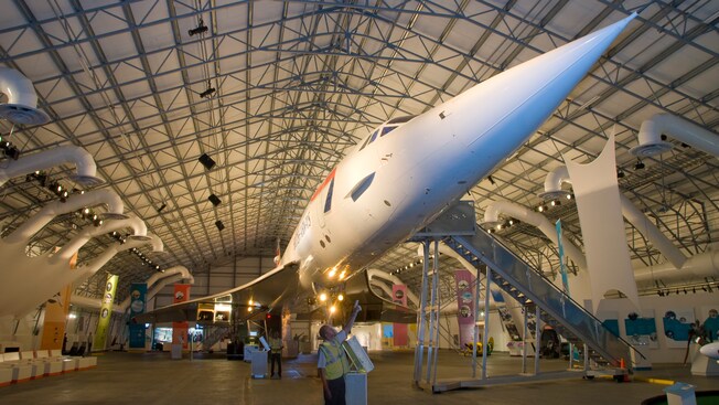 Um avião a jato da linha Concorde parado dentro de um hangar de aviões enquanto um homem aponta para seu nariz em formato de cone