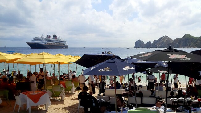 People in the ocean and sitting in chairs around tables under umbrellas on the beach with a Disney Cruise Line ship in the distance