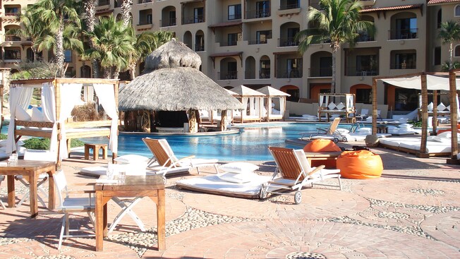 Small wooden tables and collapsible chairs and canopy beds sit on the pool deck of a hotel