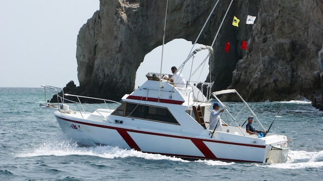 Men recline on the back of a motor boat with fishing rods as it motors past a stone arch Men recline on the back of a motor boat with fishing rods as it motors past a stone arch