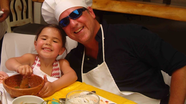 A man in an apron and chefs toque smiles alongside a young girl making salsa A man in an apron and chefs toque smiles alongside a young girl making salsa