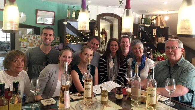 Um grupo de pessoas sorri atrás de uma mesa com garrafas de vinho, pão e pastas