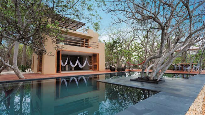 A pool in front of a 3-story pool house with tables and lounge chairs nearby