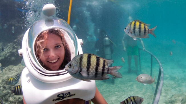 A helmet diver smiles as a fish swims in front of her underwater
