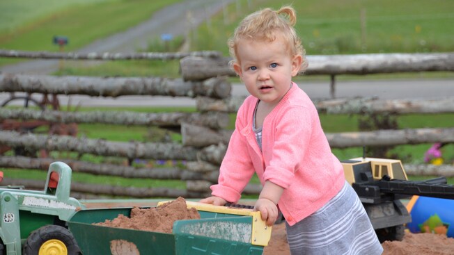 A toddler girl plays with a dump truck full of dirt near a low log fence