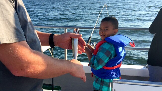 Un hombre sostiene un pez parado junto a un muchacho que pesca al costado de un bote