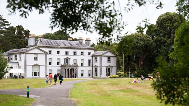 A family walks up a path to the front door of a grand 3 story mansion while people rest on the grass outside