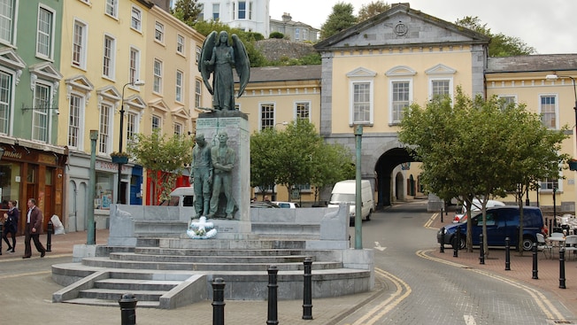 People walk near buildings and a monument with statues People walk near buildings and a monument with statues